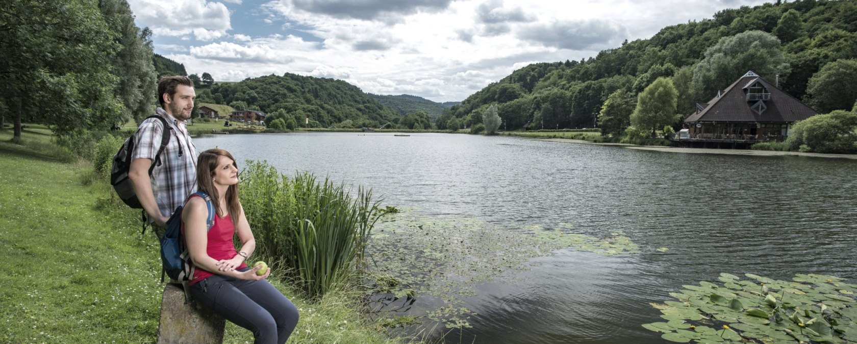 Zwei Personen sitzen am Ufer eines Sees, umgeben von grünen Hügeln und Bäumen. Ein Haus steht am rechten Ufer. Der Himmel ist bewölkt., © Kappest/REMET Zwei Personen sitzen am Ufer eines Sees, umgeben von grünen Hügeln und Bäumen. Ein Haus steht am rechten Ufer. Der Himmel ist bewölkt., © Kappest/REMET
