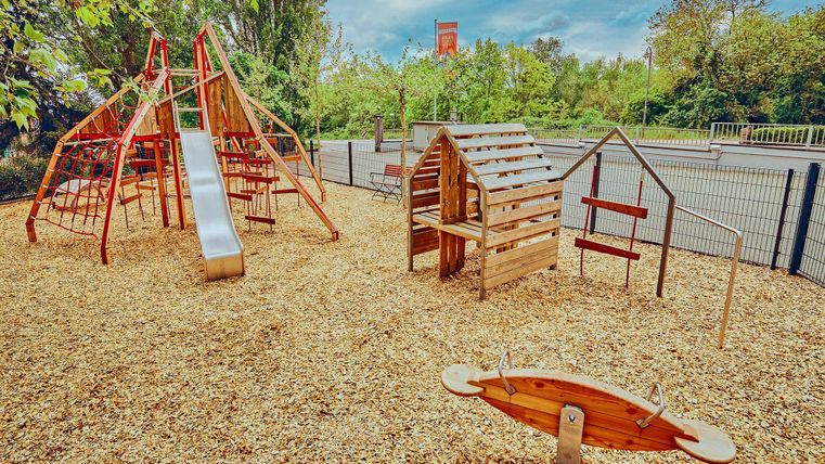 Ein moderner Spielplatz mit einer Rutsche, Klettergerüst und Holzspielhaus. Der Boden ist mit Rindenmulch bedeckt und es gibt viel Grün in der Umgebung.