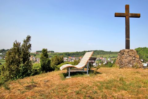 Ein ruhiger Aussichtspunkt mit einer Liege und einem Holzkreuz. Im Hintergrund sind sanfte Hügel und kleine Dörfer sichtbar.