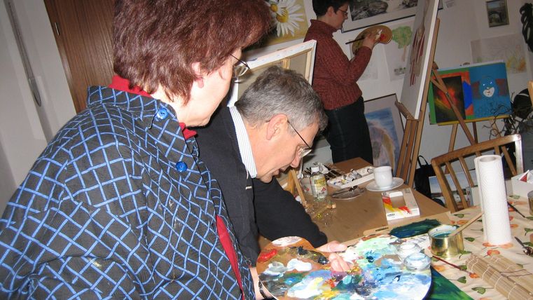 Eine Gruppe von Menschen arbeitet an ihren Malprojekten in einem hellen Raum. Pinsel und Farben sind auf dem Tisch verteilt.