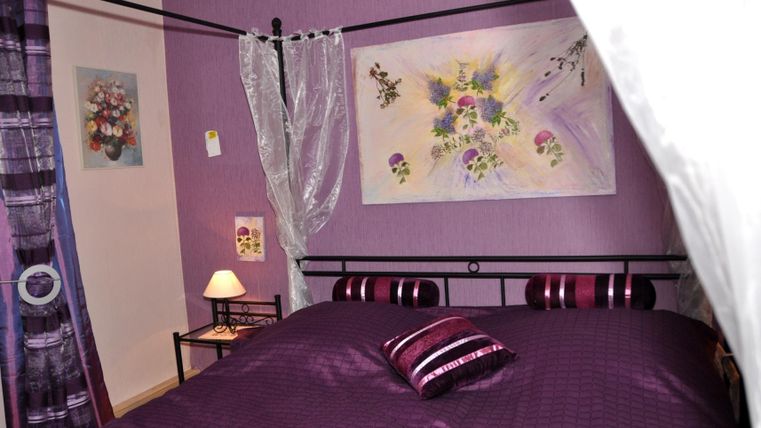 Ein gemütliches Schlafzimmer mit lila Wänden und farblich abgestimmter Bettwäsche. Ein Nachttisch mit einer Lampe und einem Gemälde an der Wand vervollständigen die Atmosphäre.