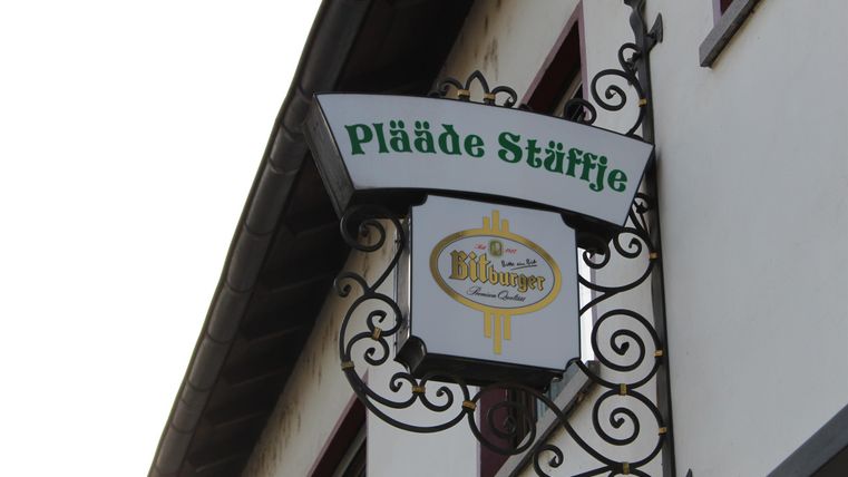 Ein hübsches Schild mit dem Namen "Pläde Stäffle" hängt an einem Gebäude. Es zeigt eine elegante Schriftart und ist mit geschwungenen Verzierungen gestaltet.