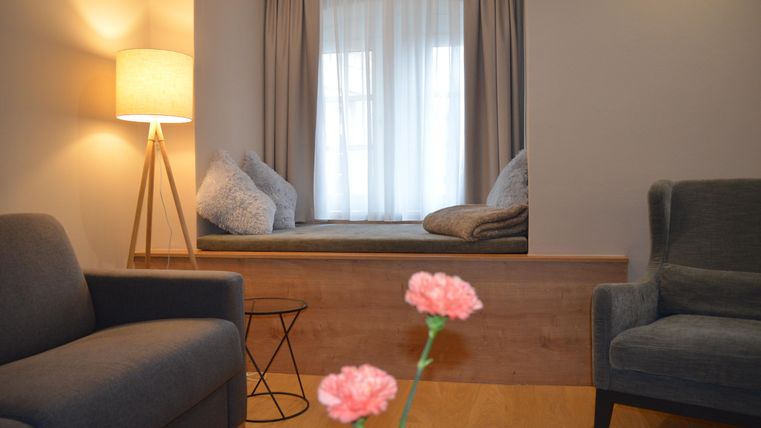 Ein gemütlicher Wohnbereich mit zwei grauen Sesseln und einer modernen Stehlampe. An einem Fenster befindet sich eine bequeme Sitzgelegenheit mit Kissen.