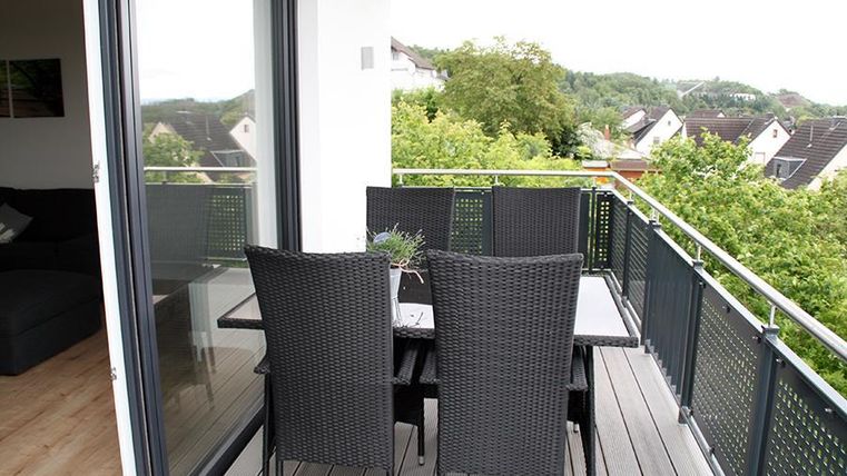 Ein moderner Balkon mit zwei schwarzen Rattanstühlen und einem Tisch. Im Hintergrund sind grüne Bäume und eine ruhige Wohngegend sichtbar.