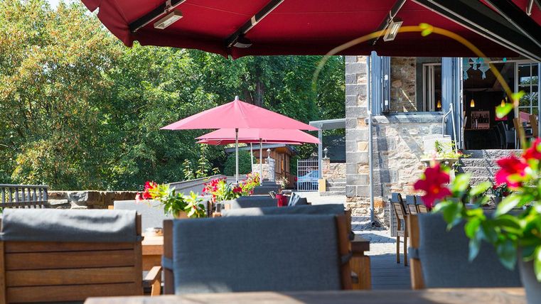 A cozy outdoor area of a restaurant with wooden tables and colorful flowers. Red umbrellas provide shade on a sunny day.