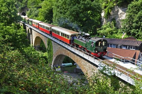 Eine Dampflok fährt über eine steinerne Brücke inmitten von grünem Wald. Die bunten Wagen der Lokomotive fügen sich harmonisch in die schöne Landschaft ein.