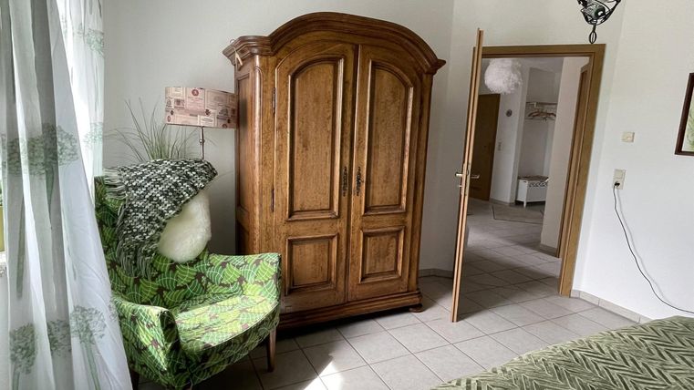 Ein gemütliches Zimmer mit einem alten Holzschrank und einem grünen Sessel. Der Raum hat große Fenster, die viel Licht hereinscheinen lassen.