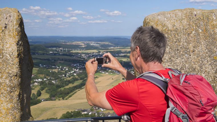Ein Mann mit einem roten T-Shirt macht ein Foto von der Aussicht. Im Hintergrund sind Landschaft und Himmel zu sehen.
