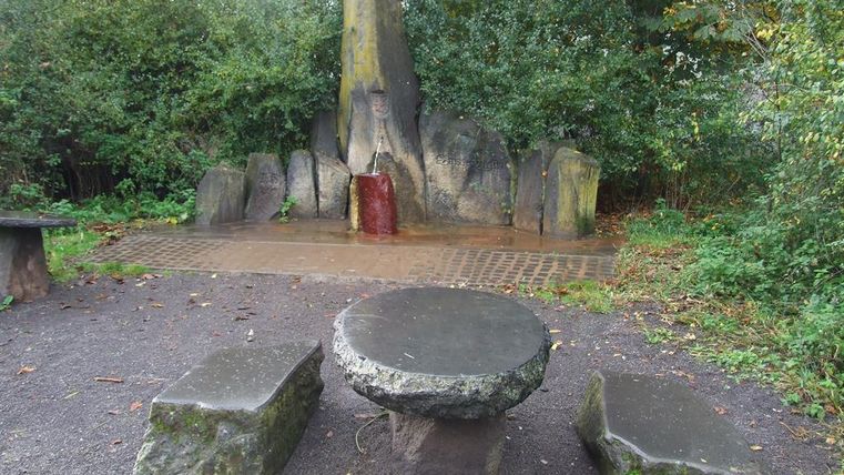 Ein Bereich im Freien mit einem runden Steintisch und zwei Sitzsteinen. Im Hintergrund steht ein großer, natürlicher Stein mit etwas rotem Wasser.