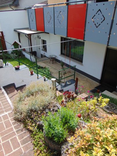 A small garden with various plants and a cozy seating area. In the background, there is a house with colorful balconies.
