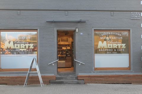Ein einladendes Café mit einer grauen Fassade und großen Fenstern. Die Fenster zeigen das Logo von "Christian Murtz Bäckerei & Café" sowie Produkte wie Brot und Kuchen.