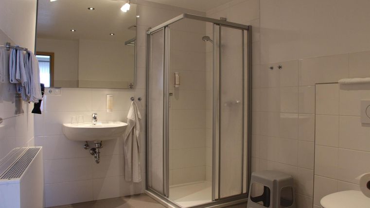 Ein modernes Badezimmer mit einer Duschkabine und einem Waschbecken. Die Wände sind in neutralen Farben gehalten und es gibt einige Handtücher und eine dekorative Sitzgelegenheit.