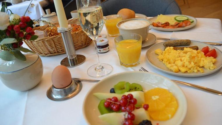 Ein reichhaltiges Frühstück mit frischem Obst, Rühreiern und Würstchen. Dazu gibt es Brötchen, Säfte und ein Glas Sekt.
