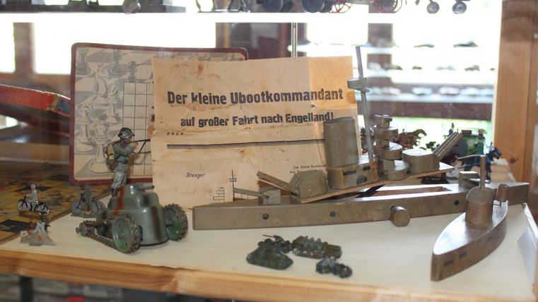 Eine Vitrine mit historischen Spielzeugen, darunter ein Modell eines U-Bootes und kleine Figuren. Dazu liegt ein Buch mit dem Titel „Der kleine Ubootkommandant auf großer Fahrt nach England“.