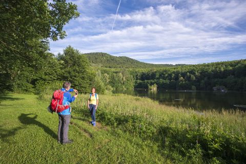 Two hikers are standing by the shore of a calm lake, surrounded by green forests and gentle hills. The sky is clear with a few clouds.