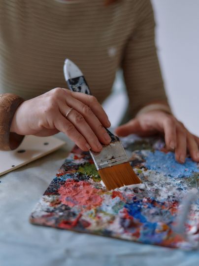 Eine Person malt mit einem Pinsel auf einer Palette voller bunter Farben. Die Hände sind konzentriert bei der Arbeit, während die Palette lebendige Farbtöne zeigt.