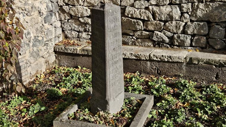 Ein grauer Obelisk steht in einem kleinen Garten, umgeben von grünen Pflanzen und Steinen. Im Hintergrund sieht man eine Mauer aus Natursteinen.