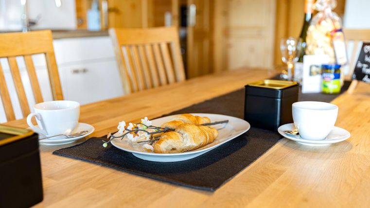 A beautifully set table with fresh pastries, a flower, and two cups. In the background, bright wooden furniture and a cozy kitchen can be seen.