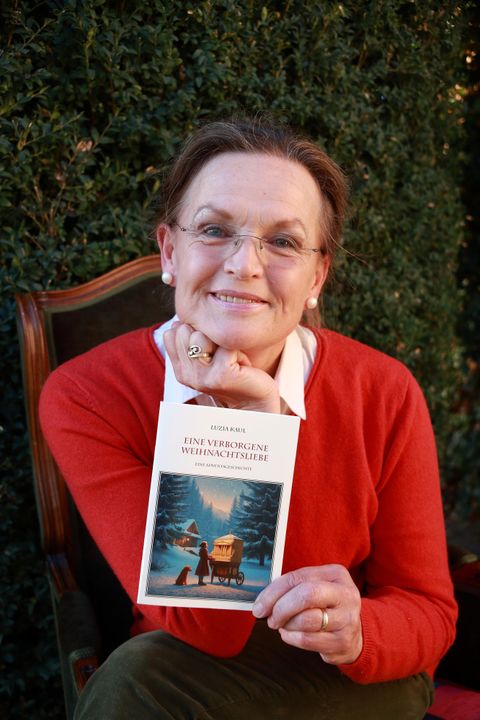 Eine lächelnde Frau hält ein Buch in der Hand und sitzt davor. Im Hintergrund sind grüne Pflanzen zu sehen.