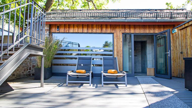 A modern terrace with two loungers and orange towels. In the background, there is a wooden house with large windows and access doors.