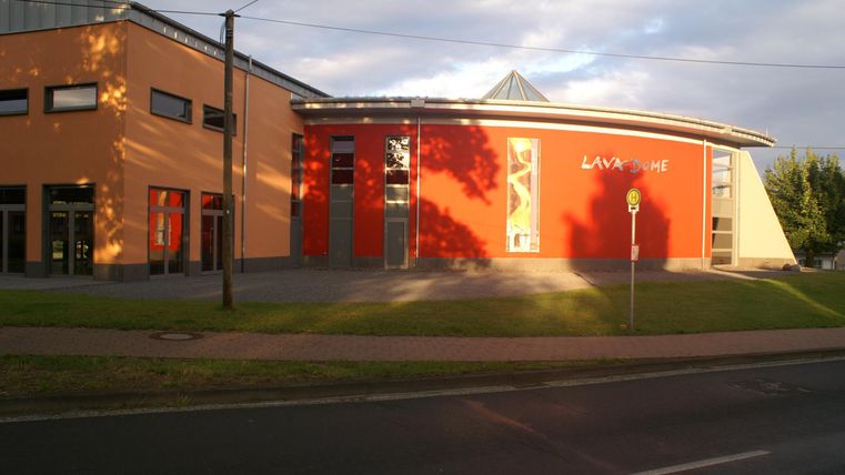 Ein modernes Gebäude mit orangefarbenen und roten Wänden. Es hat eine auffällige architektonische Form und liegt an einer Straße.