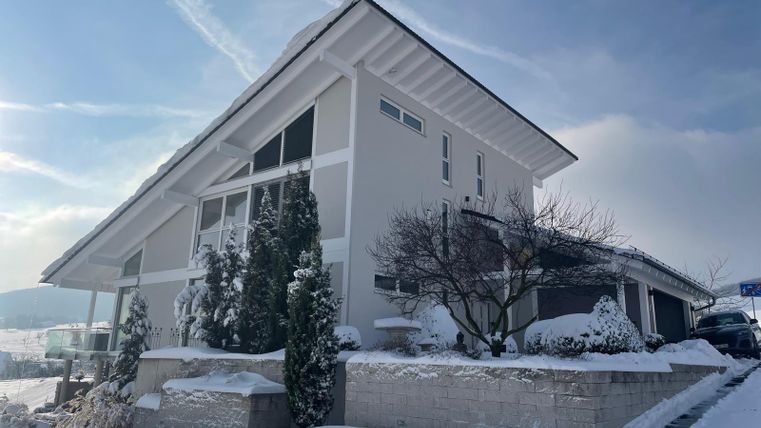 Ein modernes Haus im Schnee mit großem Fenster und umgebenden Bäumen. Der Himmel ist blau und die Landschaft ist winterlich verschneit.