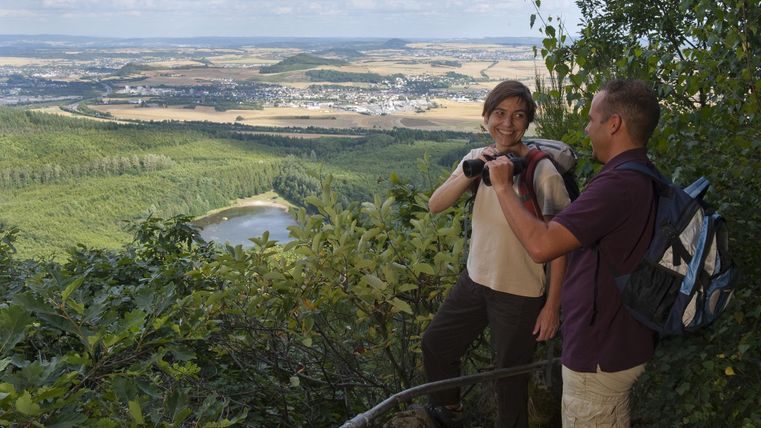 Zwei Wanderer genießen die Aussicht von einem Hügel. Im Hintergrund sind grüne Wälder und Felder zu sehen.