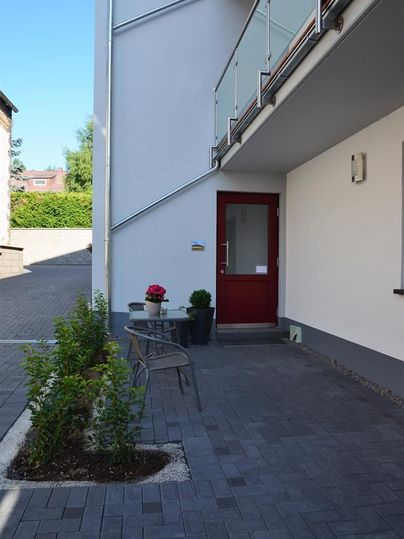 Ein moderner Eingangsbereich mit einer roten Tür und einem kleinen Sitzbereich. Umgeben von grünen Pflanzen und einem gepflasterten Weg.