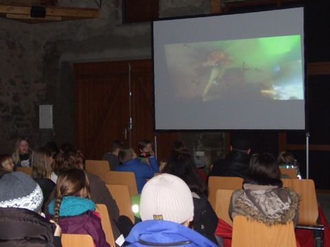 Eine Gruppe von Menschen sitzt in einem Raum und schaut sich einen Film auf einer großen Leinwand an. Der Raum hat eine rustikale Atmosphäre mit Steinen und Holz.