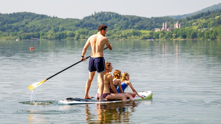 Eine Gruppe von vier Personen steht auf einem Paddleboard auf ruhigem Wasser. Im Hintergrund sind grüne Hügel und ein klarer Himmel zu sehen.