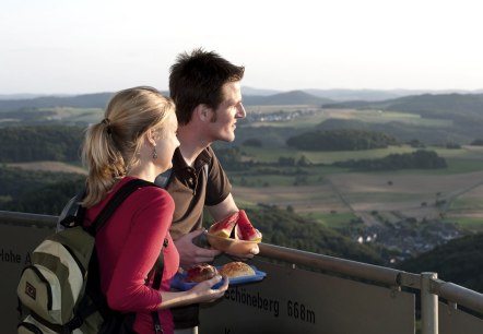 Ein Paar genie&szlig;t den Ausblick vom G&auml;nsehalsturm auf die h&uuml;gelige Landschaft. Sie halten Snacks in den H&auml;nden und schauen in die Ferne., &copy; Traumpfade