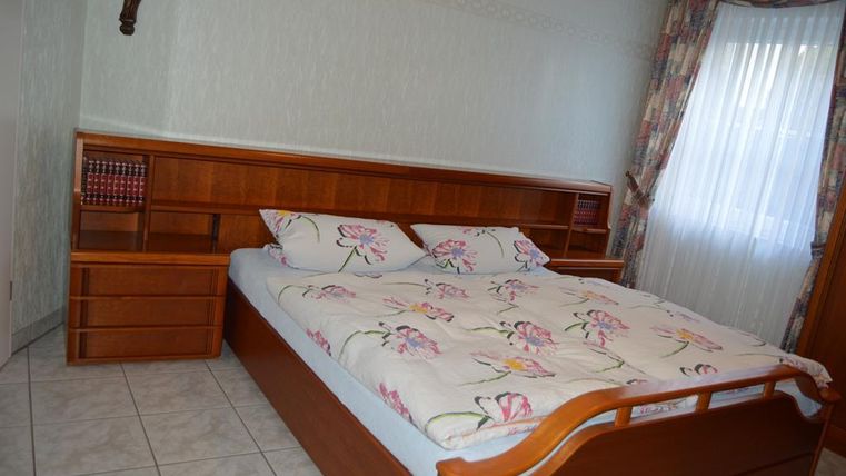 Ein einfaches Schlafzimmer mit einem großen Bett und dekorativen Kissen. Die Wände sind hell, und ein Fenster mit Vorhängen sorgt für natürliches Licht.