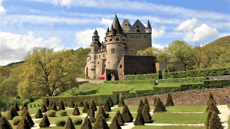 Ein beeindruckendes Schloss auf einem Hügel, umgeben von gepflegten Gärten. Die Landschaft ist grün und das Wetter ist sonnig.