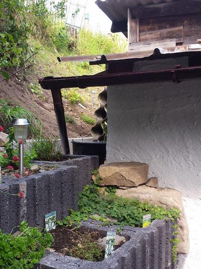 Ein kleiner Garten mit Blumenbeeten und einer Steineinfassung. Im Hintergrund steht ein Holzgebäude und die Umgebung ist grün.
