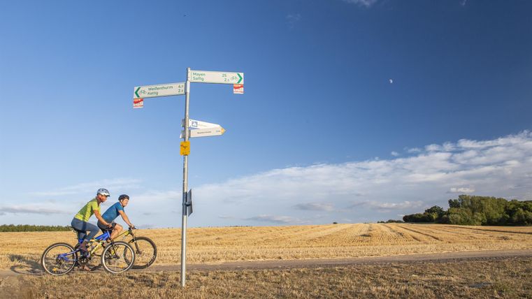 Zwei Radfahrer stehen an einem Wegweiser auf dem Land. Im Hintergrund ist ein weiter, offener Himmel zu sehen.