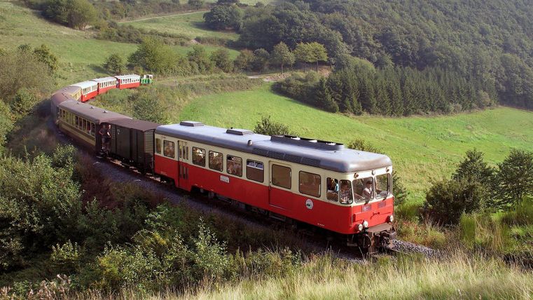 Ein roter Zug fährt durch eine grüne Hügellandschaft. Im Hintergrund sind sanfte Hügel und Bäume zu sehen.