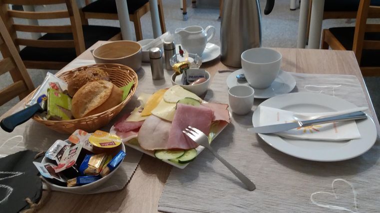 Ein gedeckter Frühstückstisch mit Brötchen, Aufschnitt und verschiedenen Marmeladen. Eine Kaffeekanne und Geschirr vervollständigen das Bild.