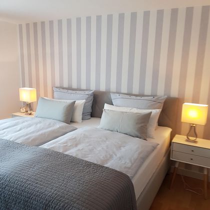 Ein gemütliches Schlafzimmer mit zwei Betten und gestreiften Wänden. Die Decke ist mit hellen Kissen dekoriert, und neben den Betten stehen stilvolle Nachttische mit Lampen.