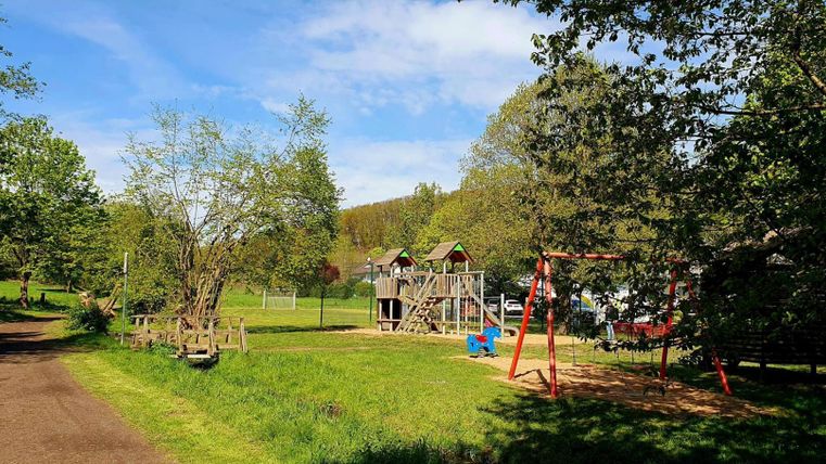 Ein schöner Spielplatz umgeben von Bäumen und Wiesen. Im Hintergrund sind eine Rutsche und Schaukeln zu sehen.
