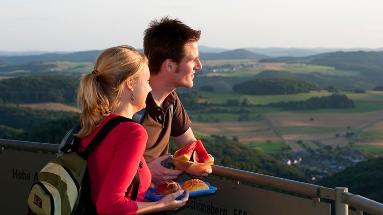 Ein Paar steht auf einem Aussichtsturm und blickt in die Ferne. Sie halten Snacks in den Händen. Im Hintergrund ist eine hügelige Landschaft zu sehen.