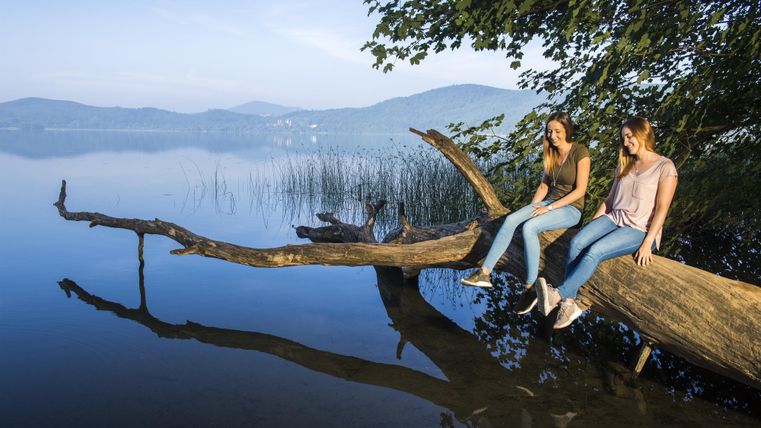 Zwei junge Frauen sitzen auf einem Baumstamm am Ufer eines ruhigen Sees. Im Hintergrund sind sanfte Hügel und ein klarer Himmel zu sehen.
