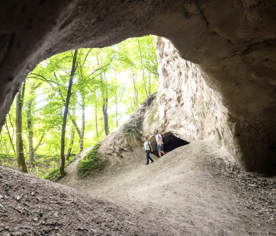 Zwei Wanderer stehen am Eingang einer Trassh&ouml;hle, umgeben von gr&uuml;nem Wald. Die H&ouml;hle ist aus hellem Gestein geformt., &copy; Eifel Tourismus GmbH, Dominik Ketz