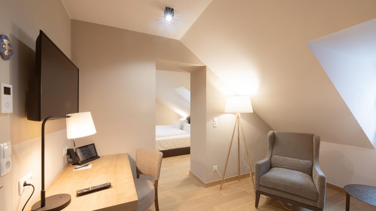 Ein modernes Zimmer mit einem TV, einem Schreibtisch und einem bequemen Sessel. Die Wände sind hell und der Boden ist aus Holz.