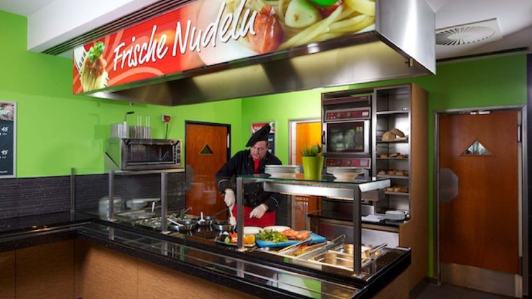 Ein modernes Nudelrestaurant mit frischem Gemüse und einem Koch hinter der Theke. Die Wände sind in einem lebhaften Grün gestrichen, und das Schild oben zeigt "Frische Nudeln".