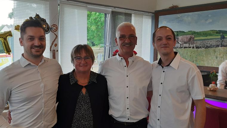 Eine Gruppe von vier Personen steht lächelnd zusammen in einem Raum. Sie tragen elegante Kleidung und wirken freundlich und einladend.