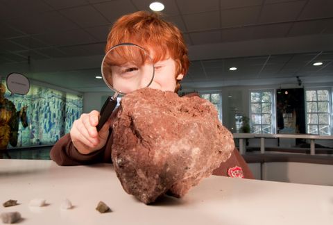 Ein Junge mit roten Haaren betrachtet einen großen Stein mit einer Lupe. Im Hintergrund sind andere Minerale und Fossilien sichtbar.