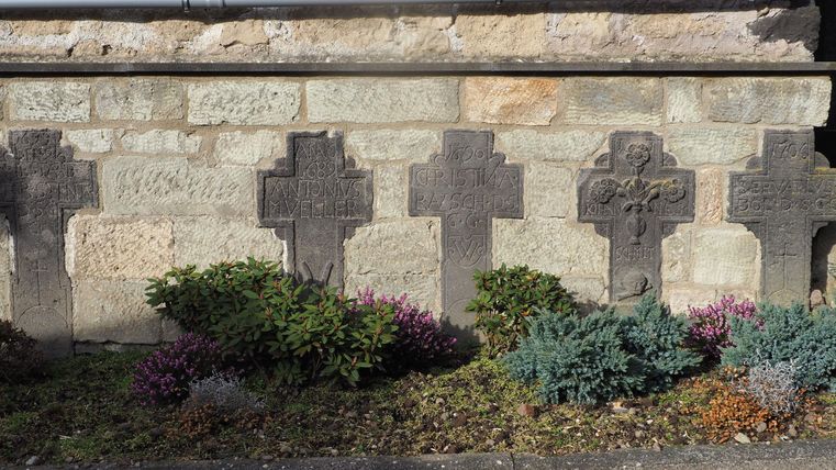 A row of stone crosses stands by a wall. Colorful plants grow in the foreground.