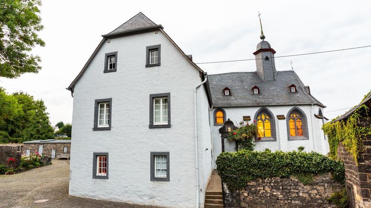 Ein historisches, weißes Gebäude mit einem grau gedeckten Dach und einem Kirchturm. Die Fenster sind hell erleuchtet und es gibt eine stimmungsvolle Atmosphäre.