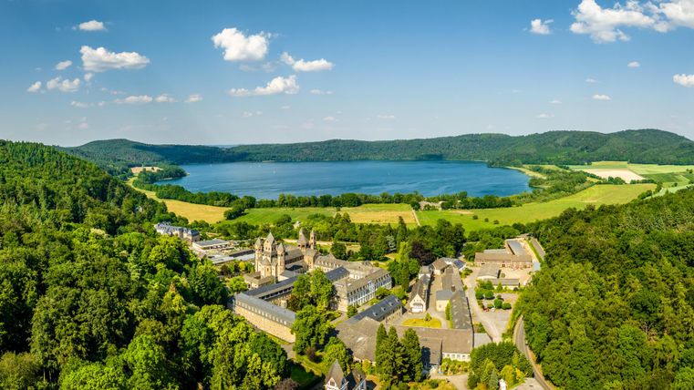 A picturesque landscape with a clear lake, surrounded by green forests and gentle hills. In the foreground, there is a historic building amidst nature.