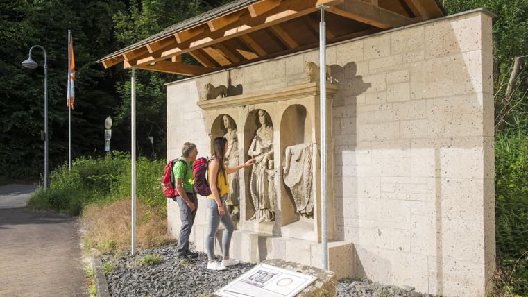 Zwei Wanderer betrachten eine kunstvolle Statue unter einem Vordach. Die Statue zeigt Figuren und ist von einem grünen Umfeld umgeben.